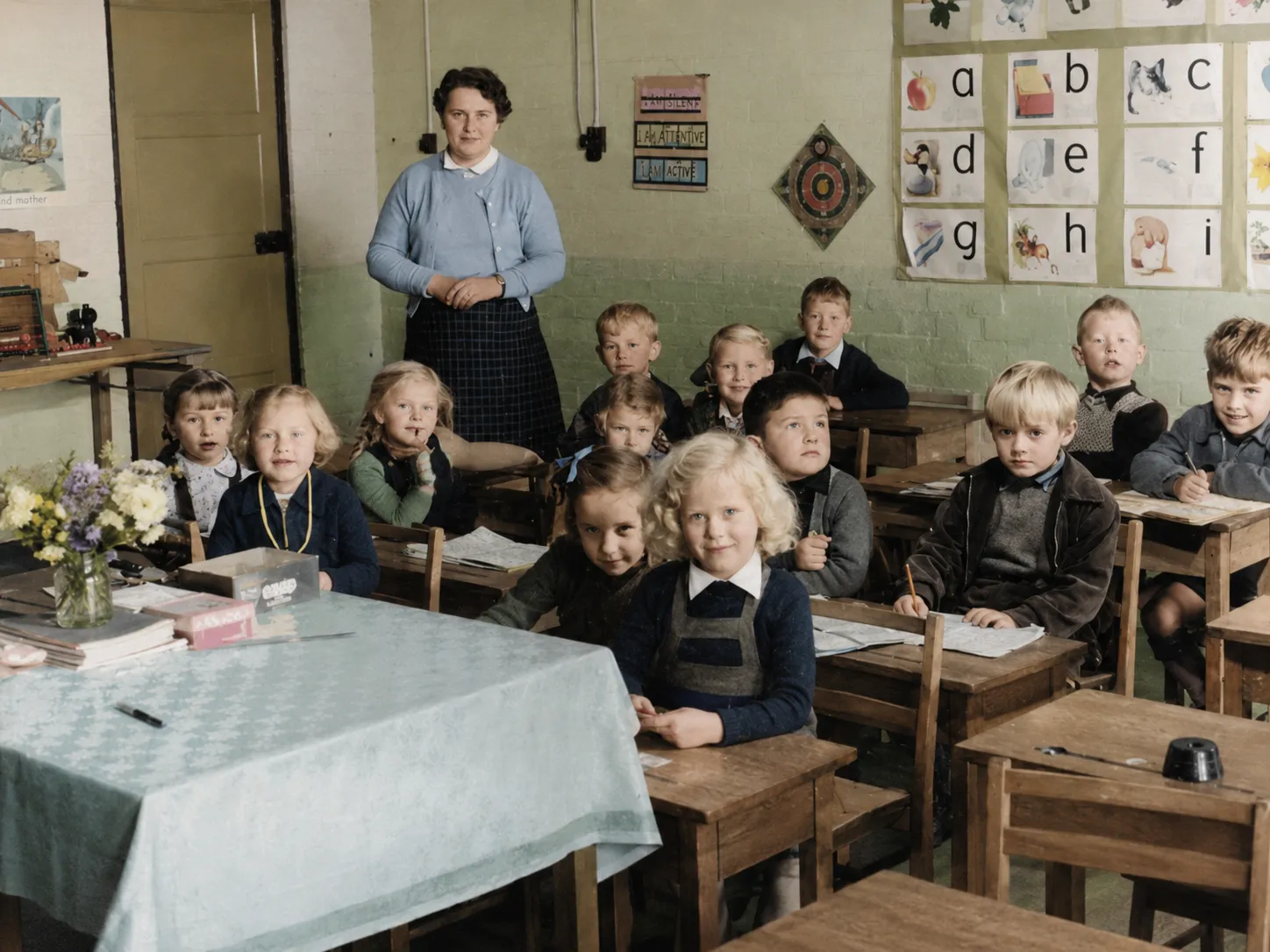 A primary-school class at the Keevil resettlement camp, Wiltshire: a teacher standing at the back of a classroom with around a dozen young children seated at wooden desks, an alphabet frieze on the wall behind them.